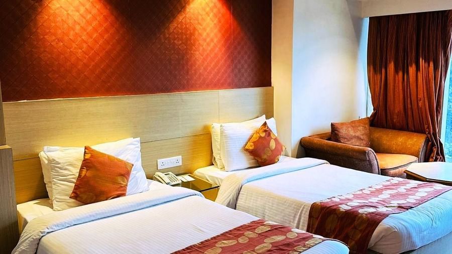 A hotel room at Amer Greens featuring two twin beds with white linen and patterned orange runners, against a textured red headboard.
