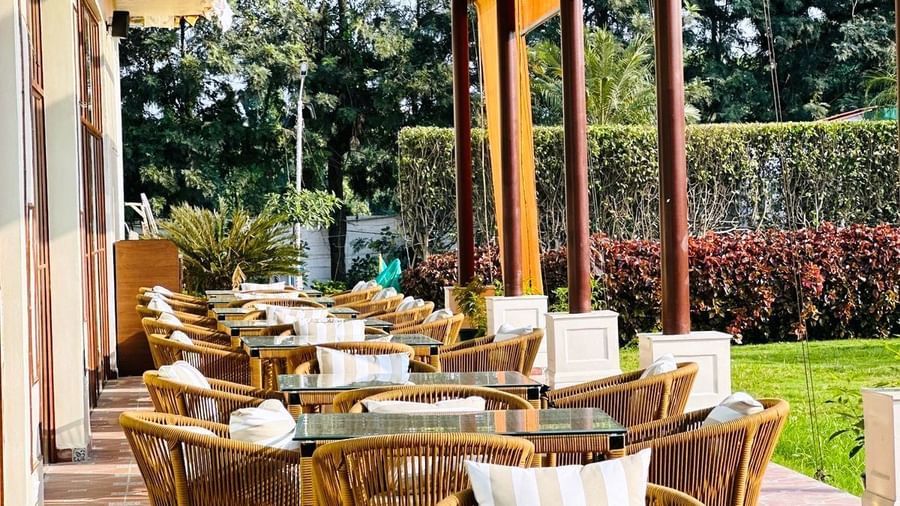  An outdoor dining terrace at Amer Greens featuring multiple woven chairs and glass-top tables under a covered pergola with a view of a lush green lawn.