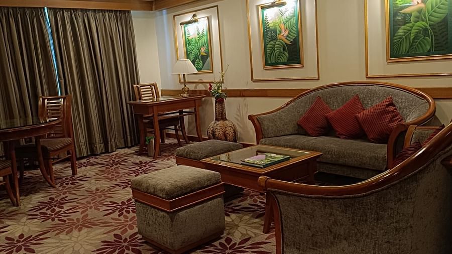 A living room featuring a floral-patterned carpet, grey upholstered seating, wooden furniture, and tropical-themed wall art with warm lighting - Pride Hotel Pune.