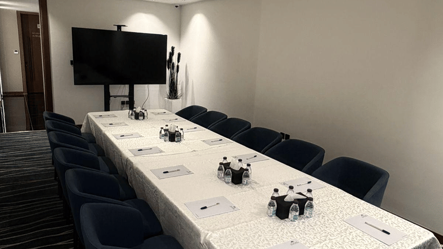 A conference boardroom at Grand Iva Hotel, Abha featuring a long table, cushioned chairs, paper pads, and a display screen. 