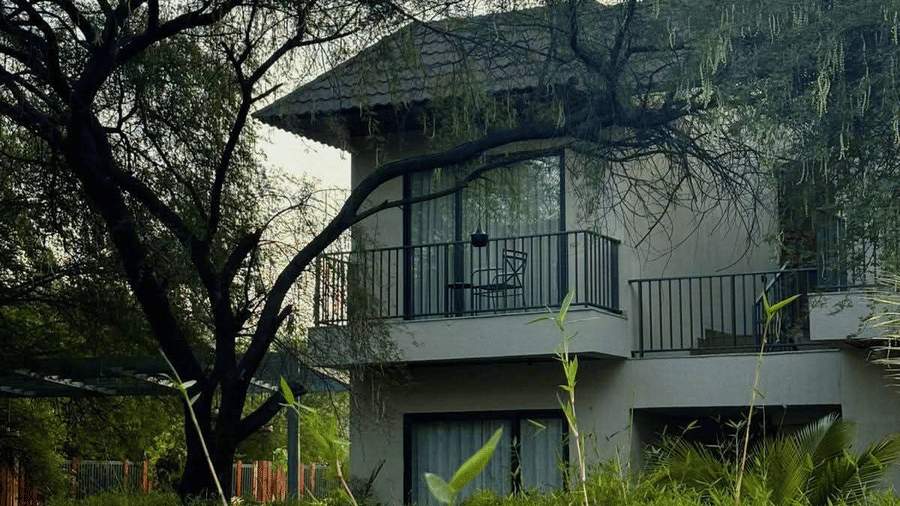 2 storey grey villa with glass windows, metal balcony railing, large tree branches, and green plants in foreground at Jivana Wellness Retreat.