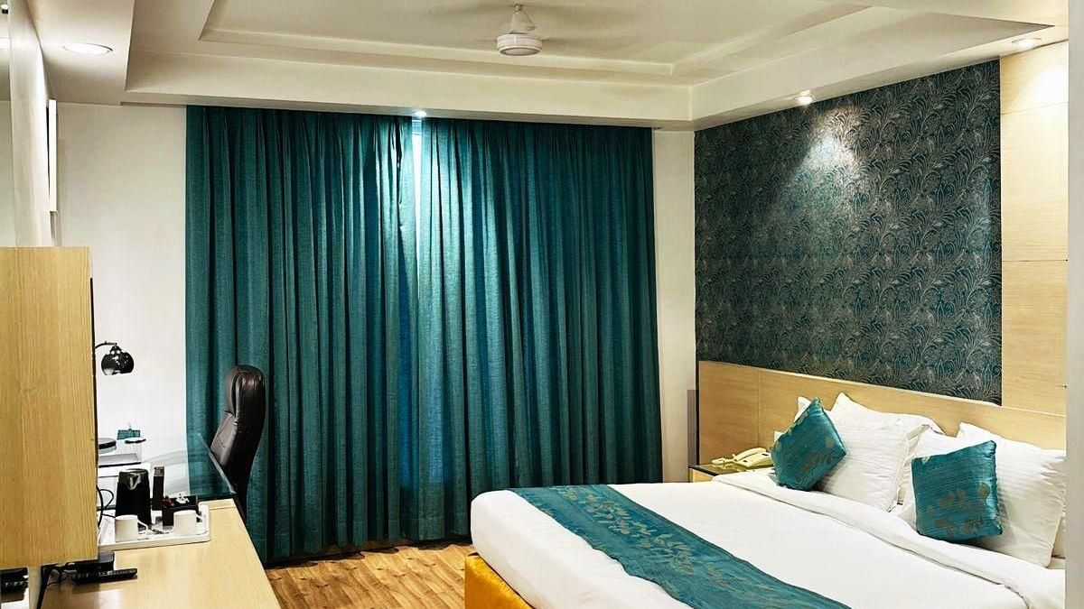 A cosy hotel room at Amer Greens featuring a large bed, hardwood floors, an accent wall, and a large window covered by floor-to-ceiling drapes. Room