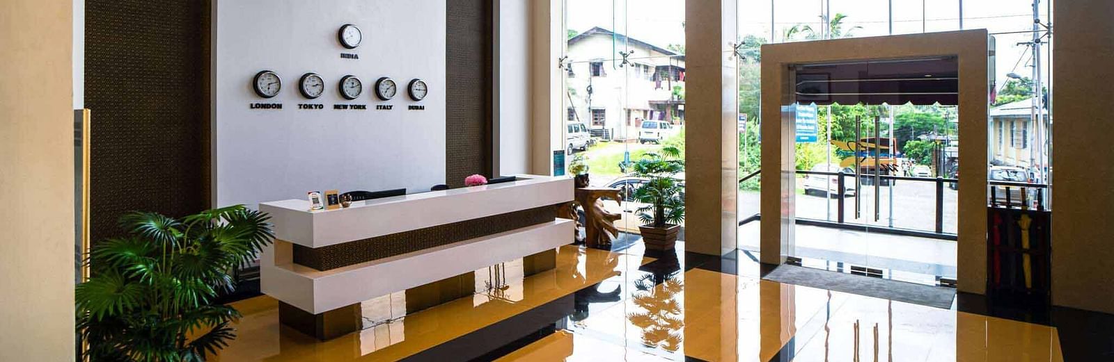 A hotel lobby at Mansha Hotel Regency featuring a white reception desk, reflective floors, and large windows with natural light overlooking the exterior with flags.