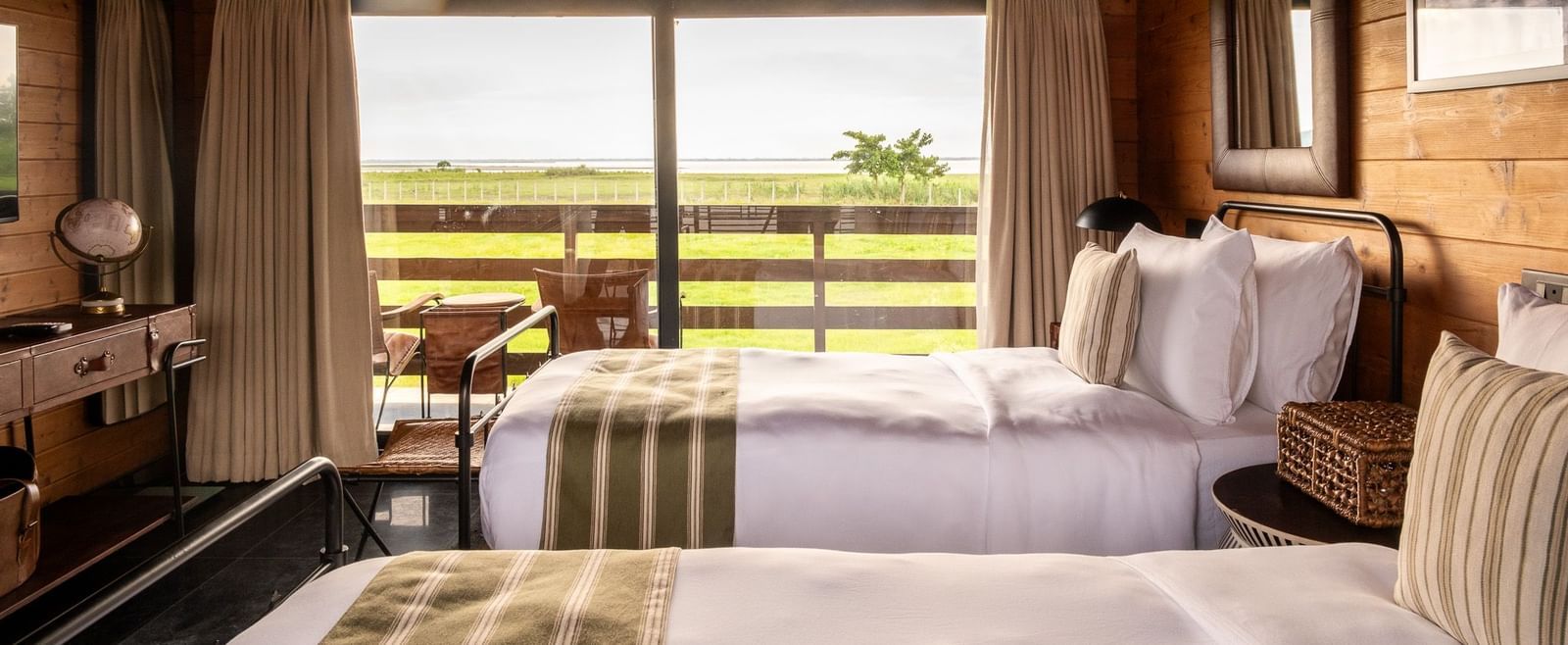 A bright and airy twin bedroom with two metal-framed beds, each with a white comforter and green striped runner. The room has wooden walls and a sloped ceiling. A large window provides a beautiful view of a grassy landscape and a body of water. This is a r