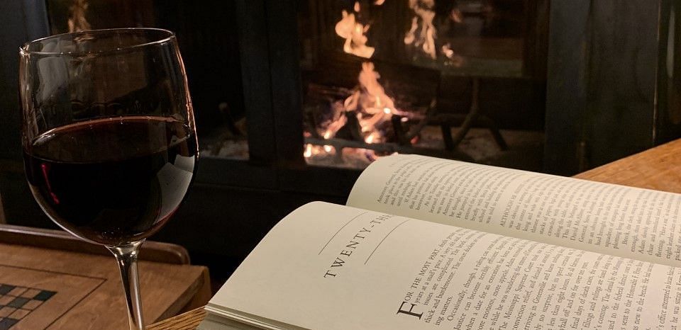 A cozy setting with an open book and a glass of wine placed on a wooden table near a fireplace, creating a warm and relaxing ambiance.