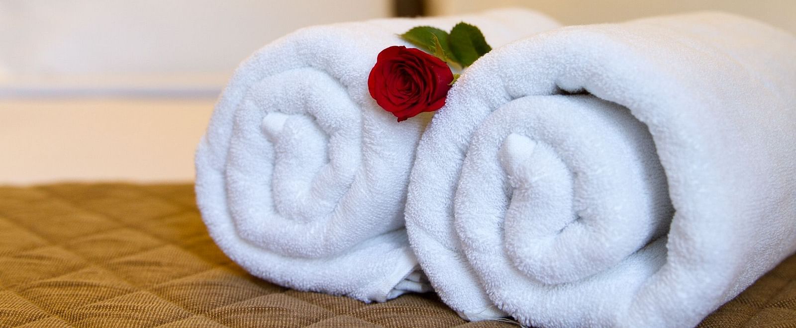 Two neatly rolled white towels are placed on a bed, adorned with a single red flower at Sanctum Suites Whitefield, Bangalore.