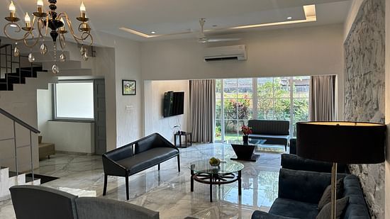 A grand living room with contemporary decor, a chandelier-lit ceiling, plush sofas, and a sleek black coffee table – Maxxvalue Klub Klass Villa, Lonavala.