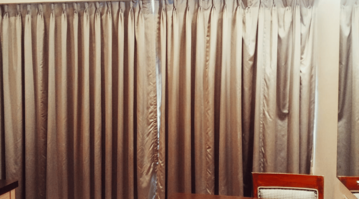 A view of a closed silver curtain next to a wooden writing desk, an office chair, and a wall-mounted air conditioner unit at Hotel Mumbai House, Thane.