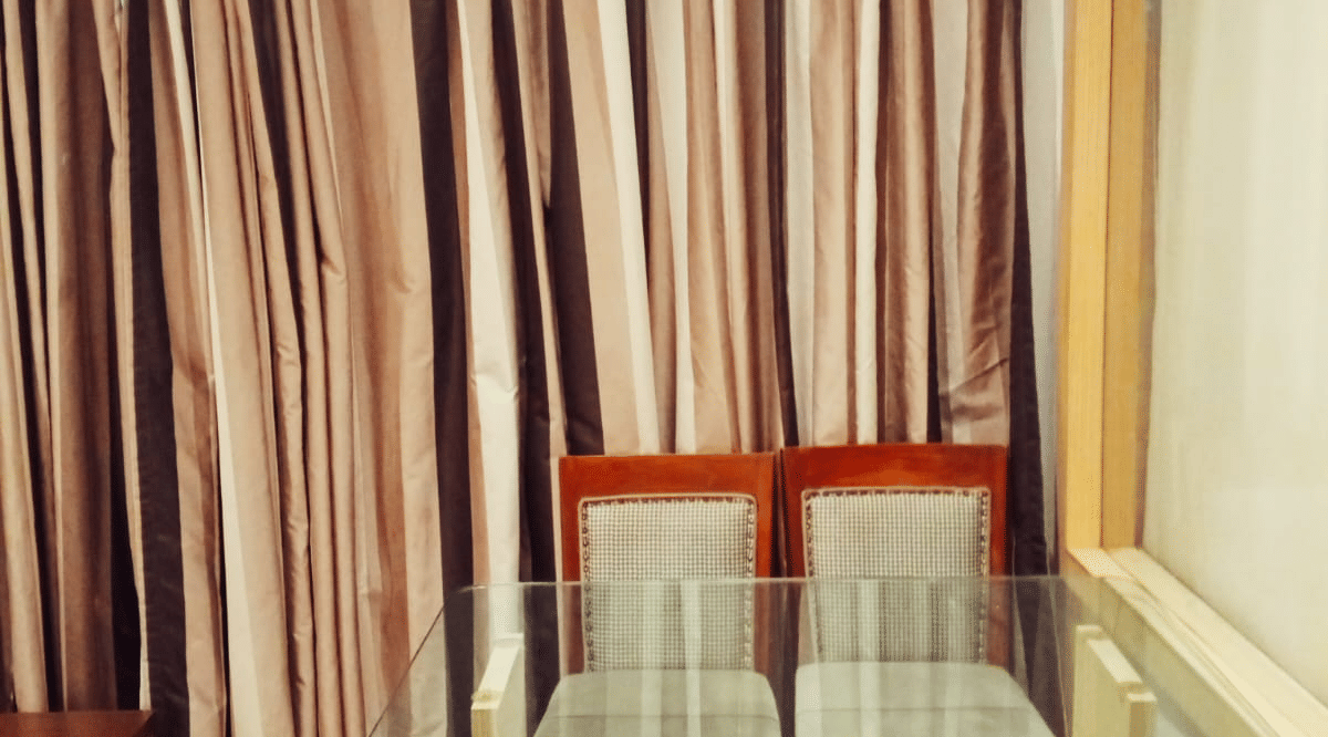 A glass top dining table paired with 4 high-back upholstered chairs positioned directly in front of floor-length curtains at Hotel Mumbai House, Thane.