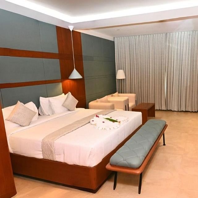 A hotel room at Dhanalakshmi Srinivasan Hotels, Perambalur, featuring a double bed, two benches, a TV on a console, a desk with a chair, two armchairs, and closed curtains.