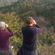2 people standing on a hill and looking through binoculars at a lush green mountain range.