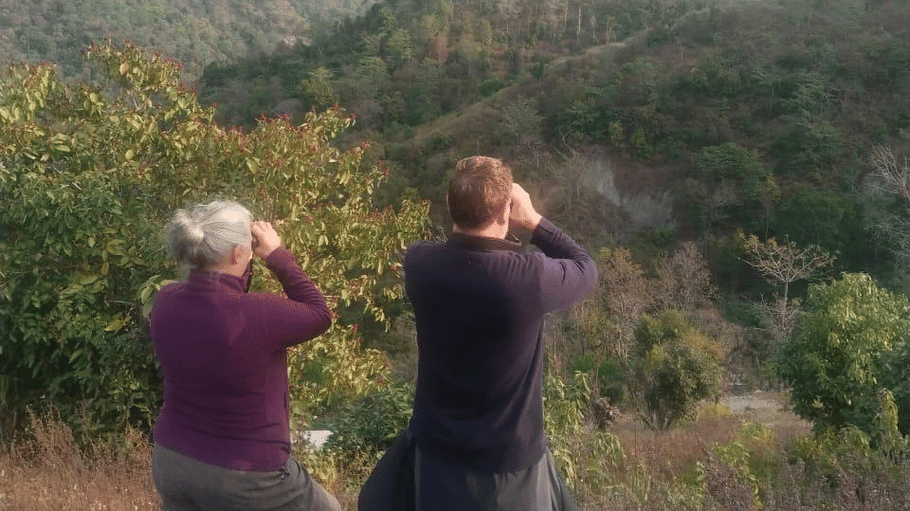 2 people standing on a hill and looking through binoculars at a lush green mountain range.