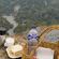 A picnic basket, flask, 1 bottle and snacks resting on a hill overlooking lush mountains.