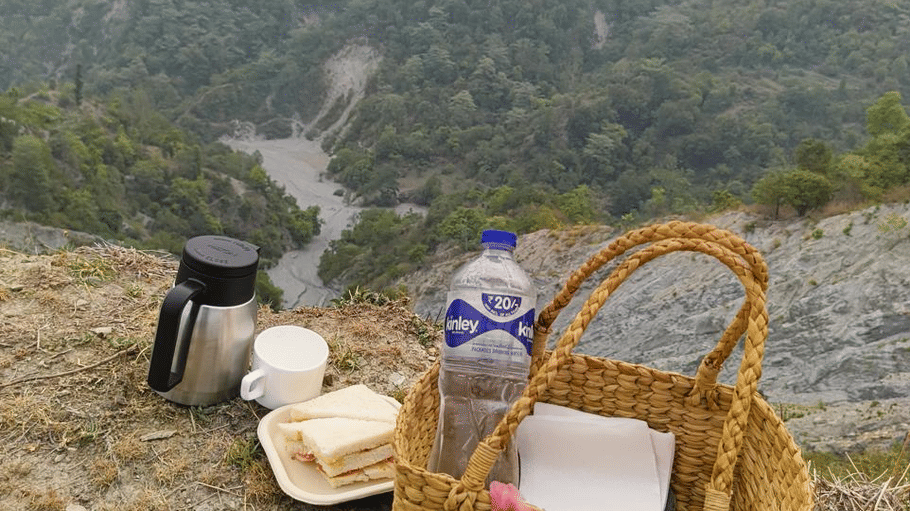 A picnic basket, flask, 1 bottle and snacks resting on a hill overlooking lush mountains.