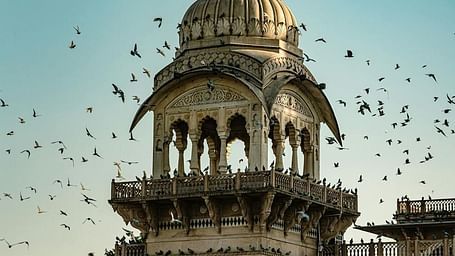 A domed tower with arched openings is part of a larger structure under a clear sky with flying birds.