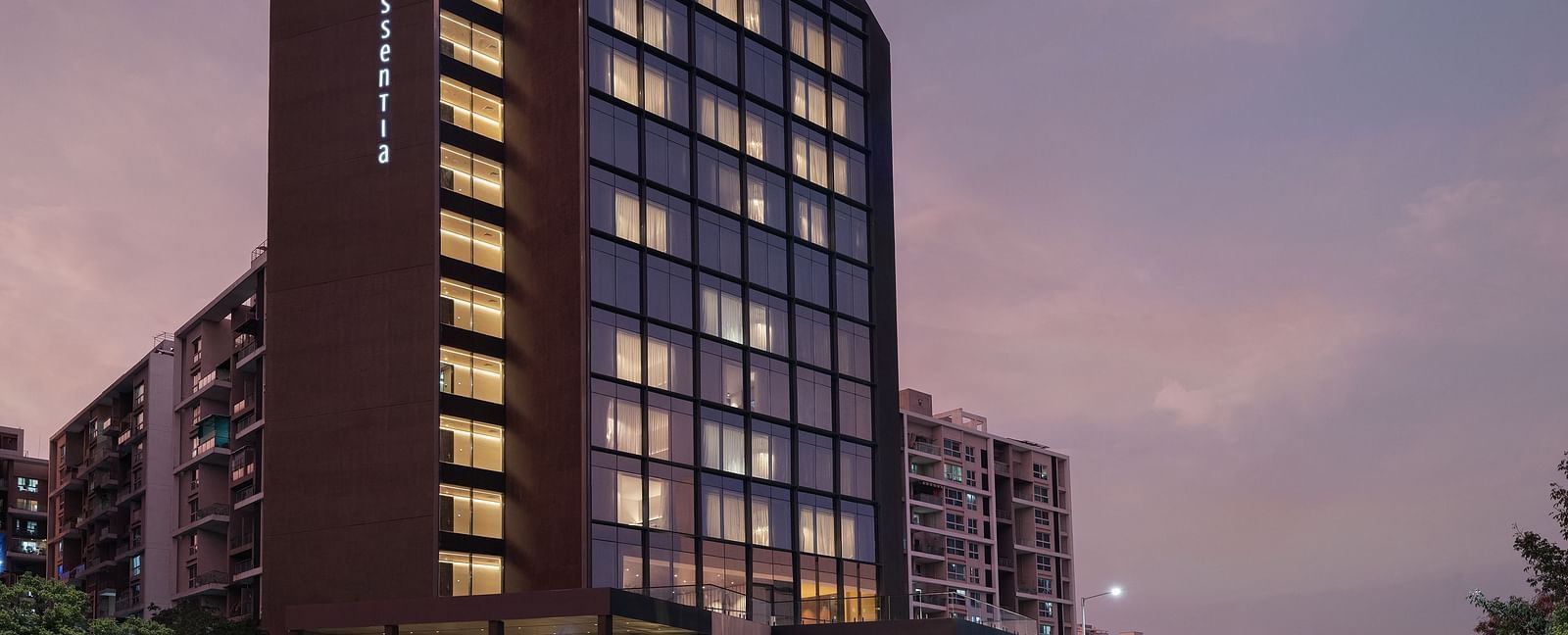 Facade of Essentia Premiere, Pune with lights on during twilight.
