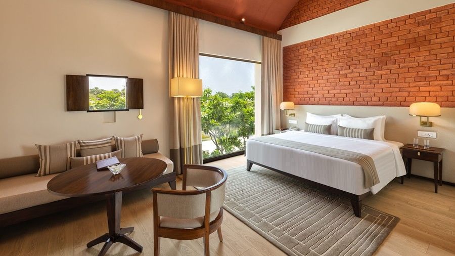 Modern hotel room with a cosy double bed, brick accent wall, large window overlooking greenery, a wooden dining table with chairs, and a sofa with striped cushions - Marasa Sarovar Premiere, Bodhgaya