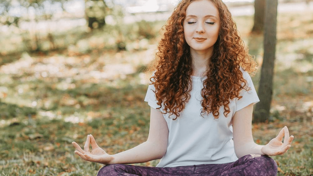 A woman meditating in a open space sitting on a mat with trees in the background.
