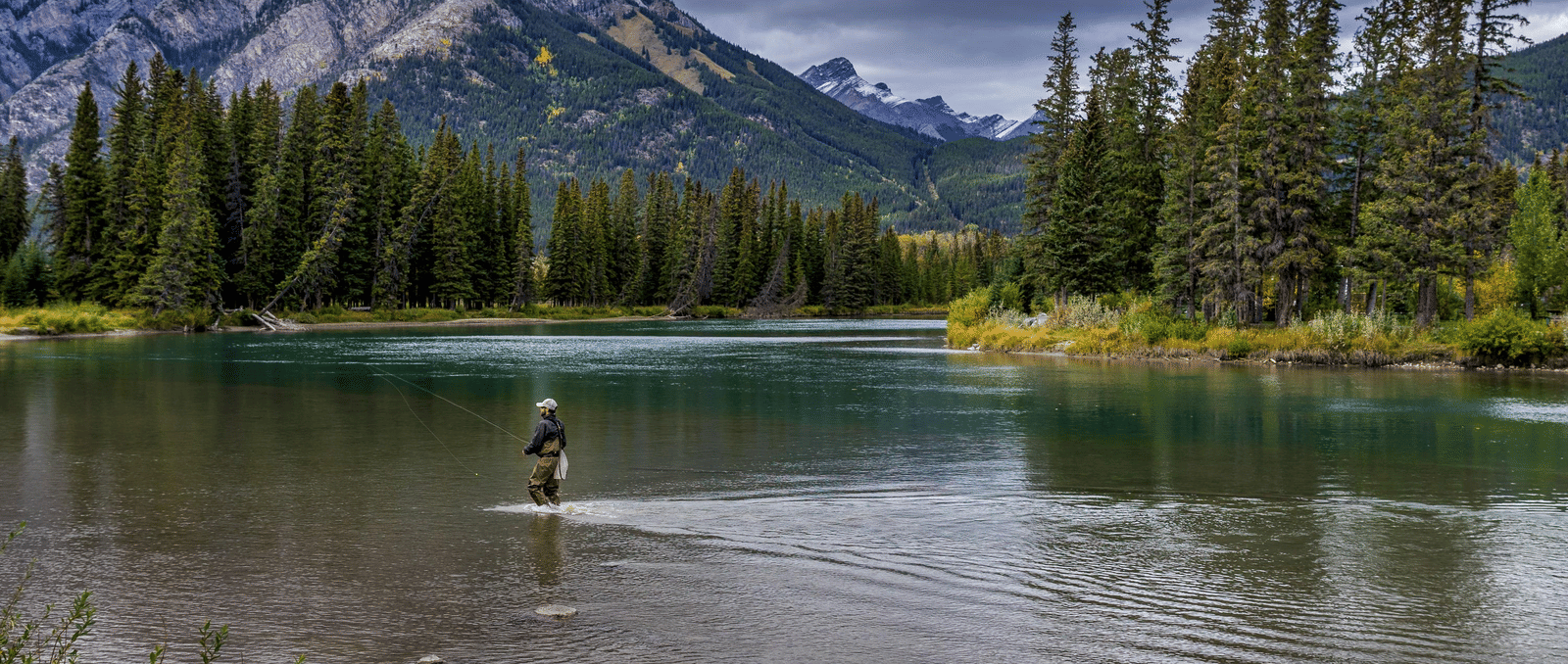 A lone person fishing in a river surrounded by greenery and a mountain.