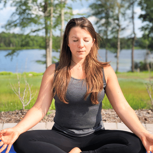Woman meditating outdoors on a blue yoga mat in a cross-legged pose, with eyes closed and a serene lake view behind her