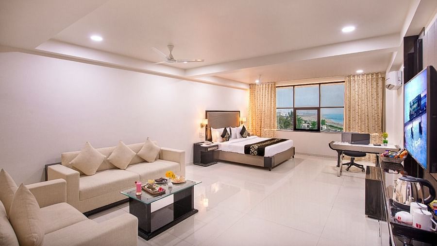 An airy hotel suite provides a comfortable seating arrangement, a television for entertainment, and expansive windows revealing an outdoor view.