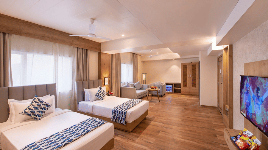 A spacious, brightly lit hotel room in Lords Inn Jamnagar, featuring wall-to-wall windows, two neatly made double beds, a wall-mounted TV, a snack-filled tray, and polished wooden flooring.