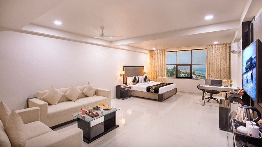 An airy hotel suite provides a comfortable seating arrangement, a television for entertainment, and expansive windows revealing an outdoor view.