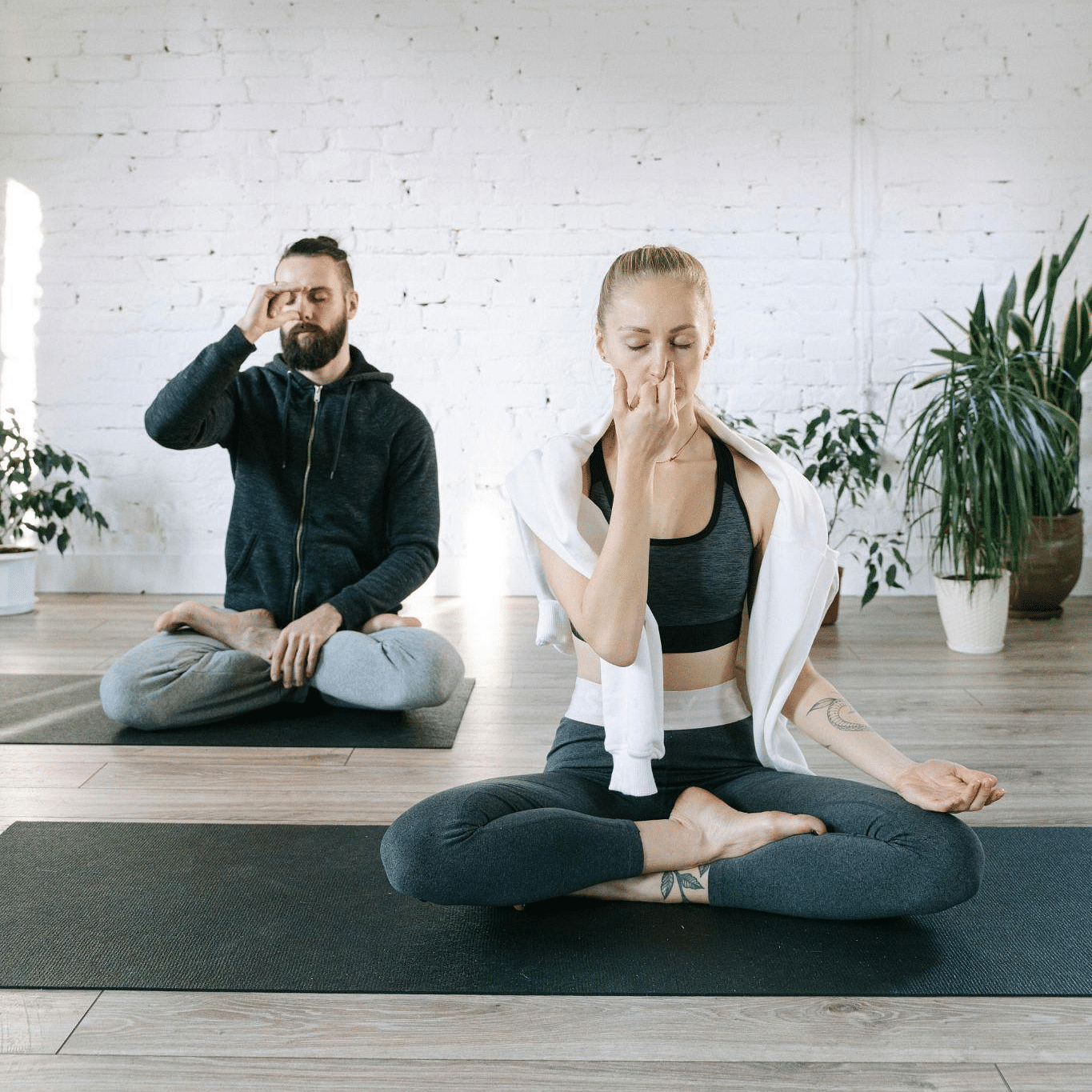 A man and woman practice alternate nostril breathing in a yoga studio, seated on mats with eyes closed.