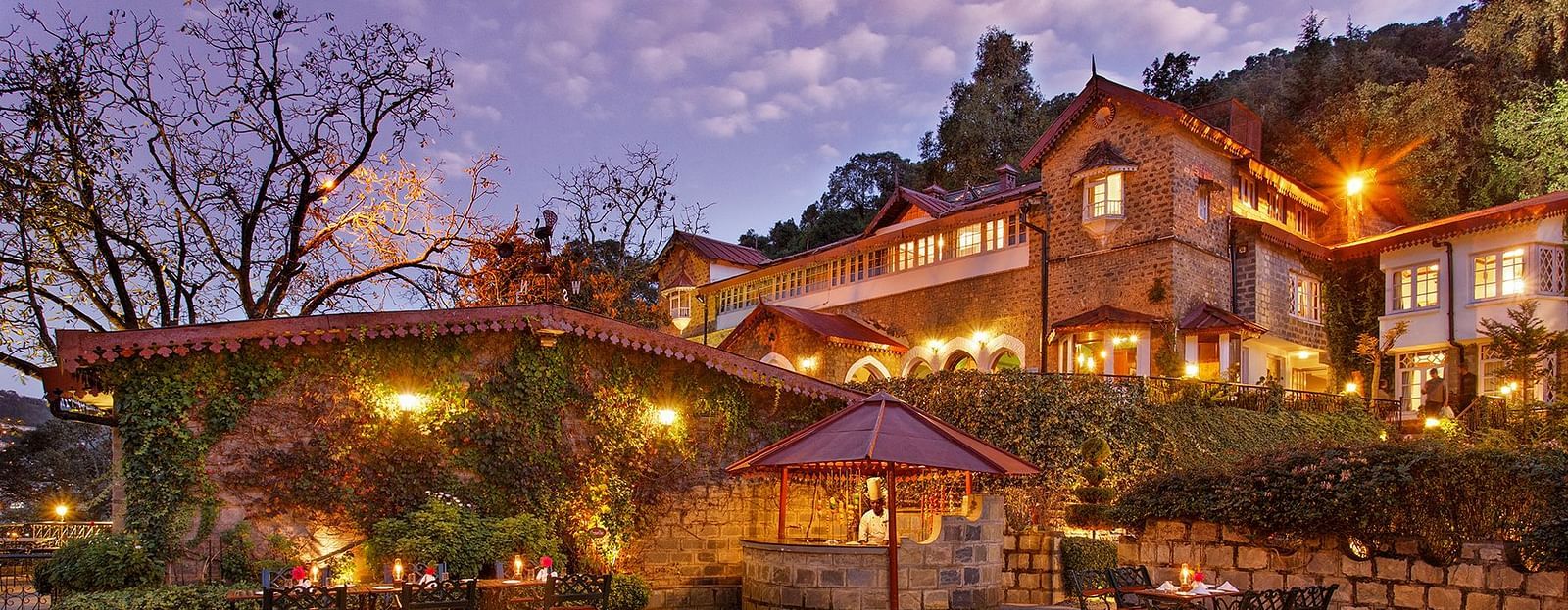 A beautiful outdoor dining area at the naini retreat, nainital. There are tables and chairs set up on a stone patio, with a fireplace in the center. The building is lit up at dusk, creating a warm atmosphere. 