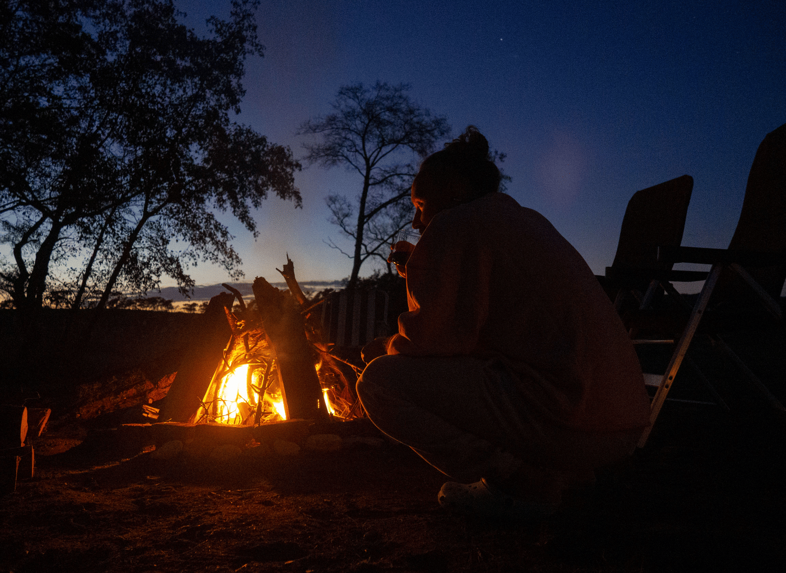 An evening gathering showing a person sitting outdoors near a large campfire under a night sky with trees.
