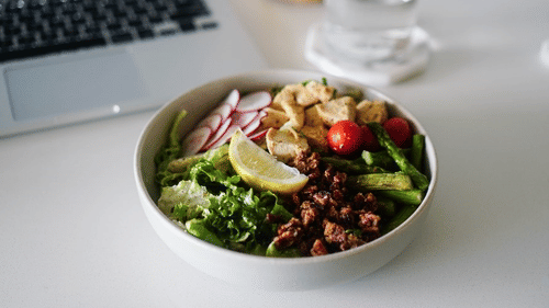 a bowl of healthy food including vegetables kept on a surface next to a laptop and a glass of water on a coaster