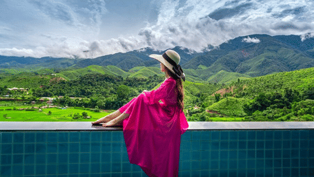 A woman in a pink dress and hat sits at the edge of an infinity pool.
