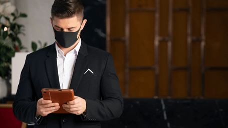 A man in a black mask and a suit holds a tablet and looks down.