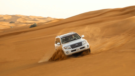 A white 4x4 car is driving up a steep sand dune, with sand spraying from its tyres.