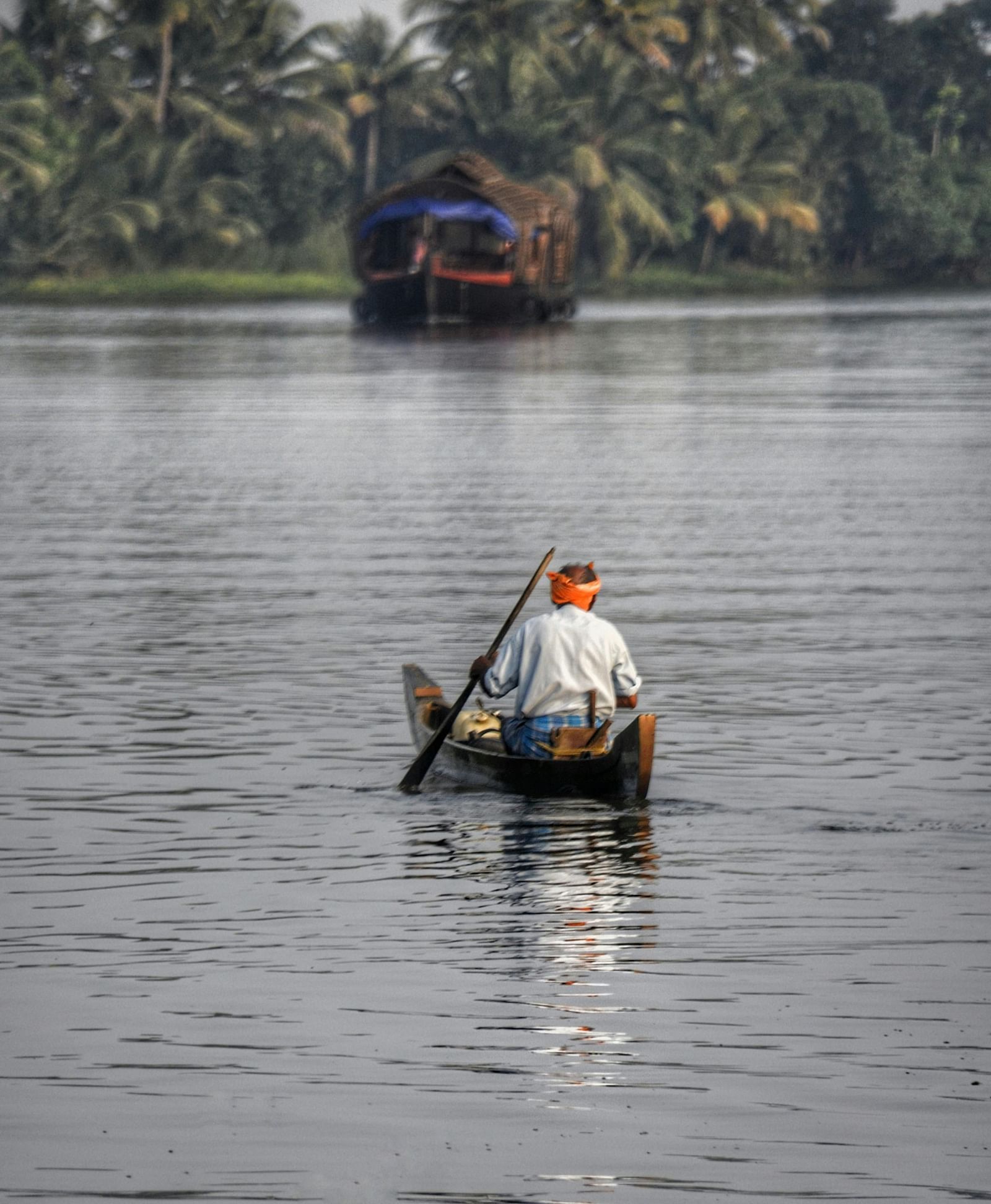 A lone fisherman rows a small boat on a tranquil river with a houseboat and lush greenery in the background