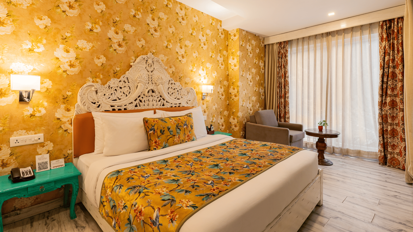 A spacious bedroom featuring a bed with a decorated headboard, an accent wall with floral wallpaper, a bedside table, and a single-seater sofa facing a small round table | Summit by the Ganges Beach Resort & Spa, Rishikesh