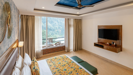 A spacious room featuring a bed facing a television unit, and an attached balcony with a 2-seater round table and 2 chairs, overlooking the trees and a river in the distance | Summit by the Ganges Beach Resort & Spa, Rishikesh