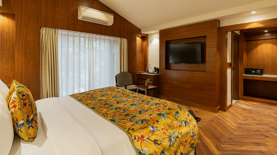 A spacious bedroom featuring a bed, an A/C unit, walls with wooden panels, a television facing the bed, and an attached bathroom | Summit by the Ganges Beach Resort & Spa, Rishikesh