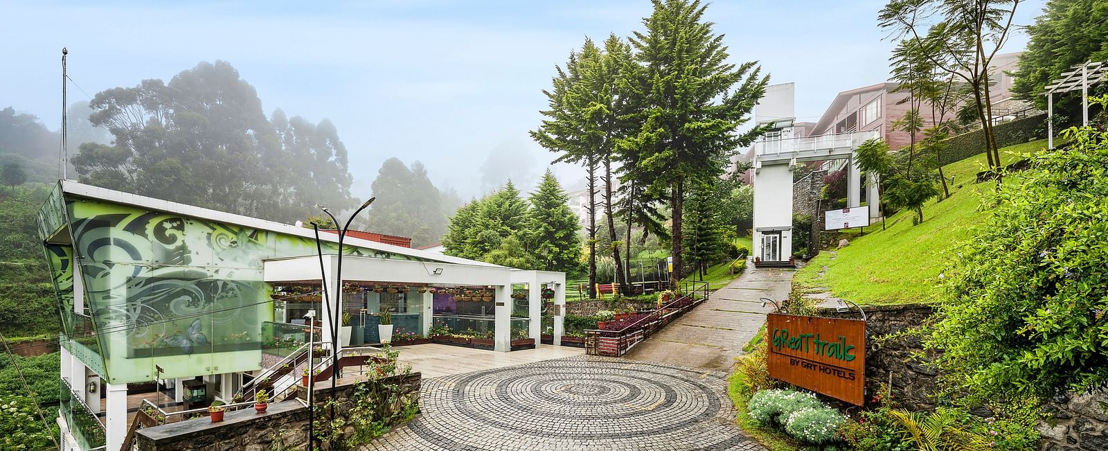 A facade image of the GReaT Trails Kodaikanal By GRT Hotels with lush greenery and the trails leading to the best mountain view resort, Kodaikanal, Tamil Nadu