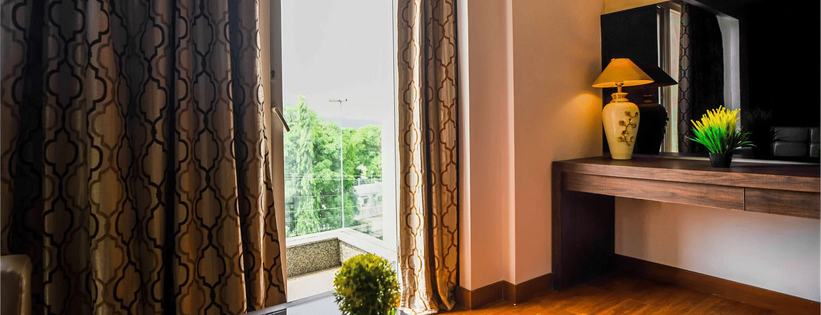 A hotel room at Mansha Hotel Regency with a large window draped with dark curtains, a wooden table, a television, with a plant on the table, and a doorway leading to the balcony.