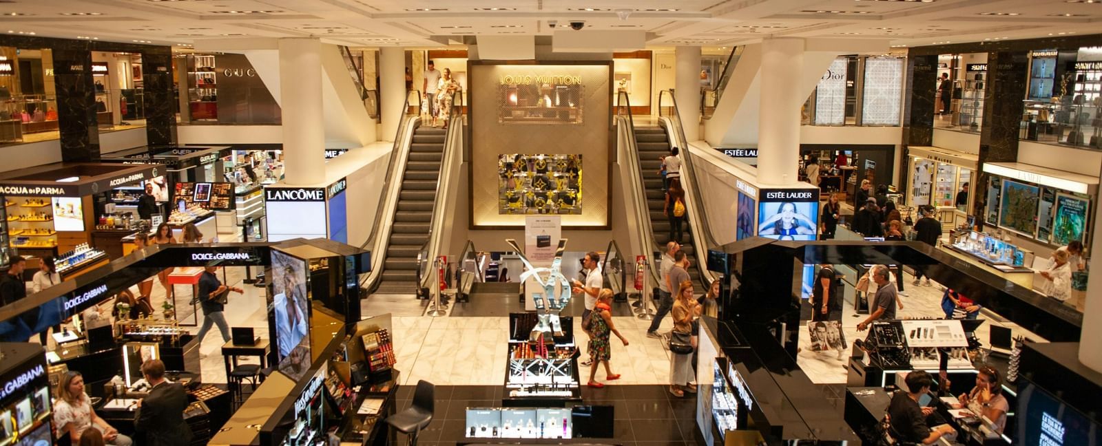 A bustling luxury shopping floor with bright displays, escalators, and well-arranged counters creating a lively retail atmosphere.