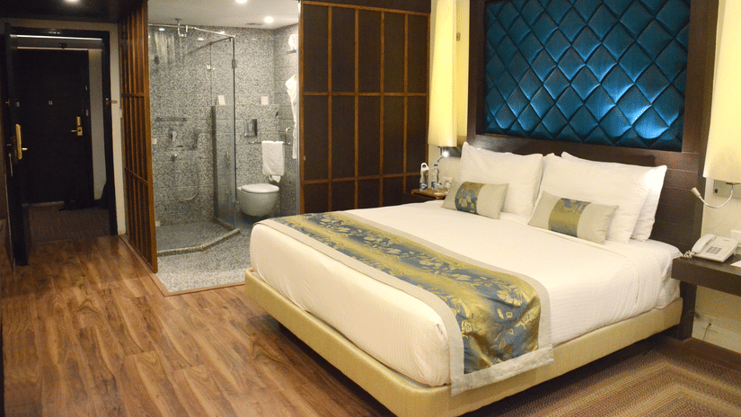 A room with a neatly made bed and bedside tables, warm lighting, and a doorway leading to a modern washroom at Clarks Avadh, Lucknow | Rooms with Gomti River View.