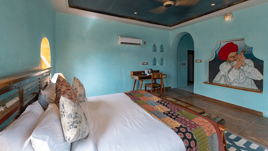 An image of a bedroom with double size bed, bedside table on either side, two open windows, and a table on one corner of the room to place various items - Karwaan Jaisalmer