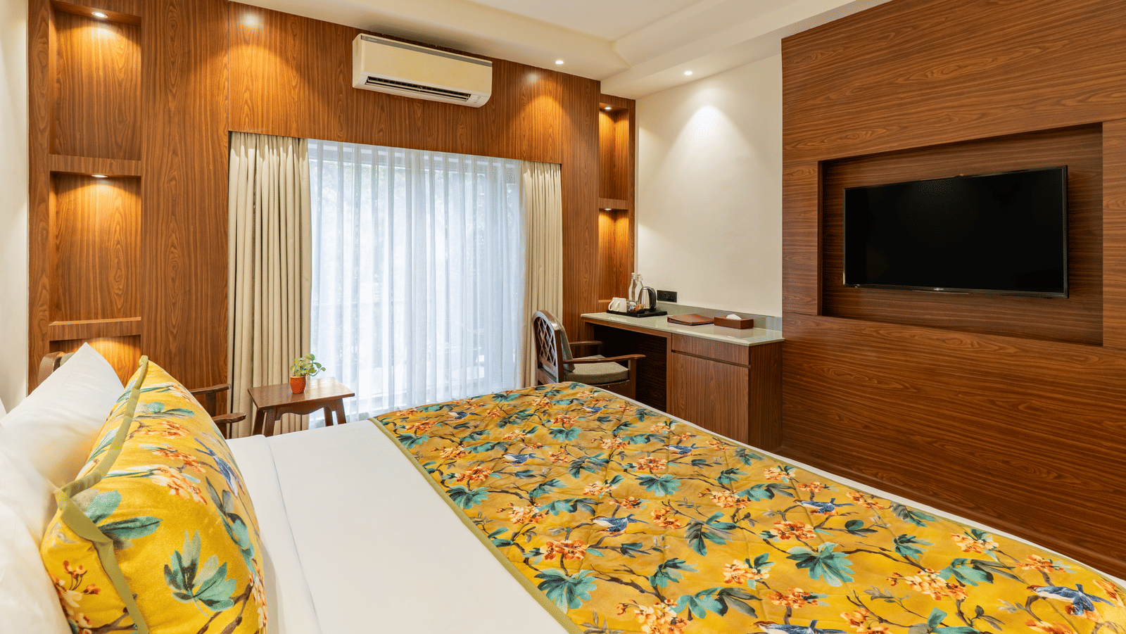 A well-lit bedroom featuring a neatly made bed facing a television unit, walls with wood panels, an A/C unit and a fan, a chair and a cabinet, and full-length curtains | Summit by the Ganges Beach Resort & Spa, Rishikesh