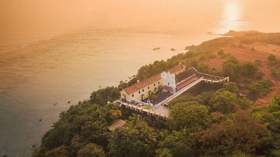 An aerial or high-angle view of a green, tree-covered headland with a building, likely Fort Tiracol, Goa, overlooking a wide body of water bathed in the warm glow of sunset or sunrise.