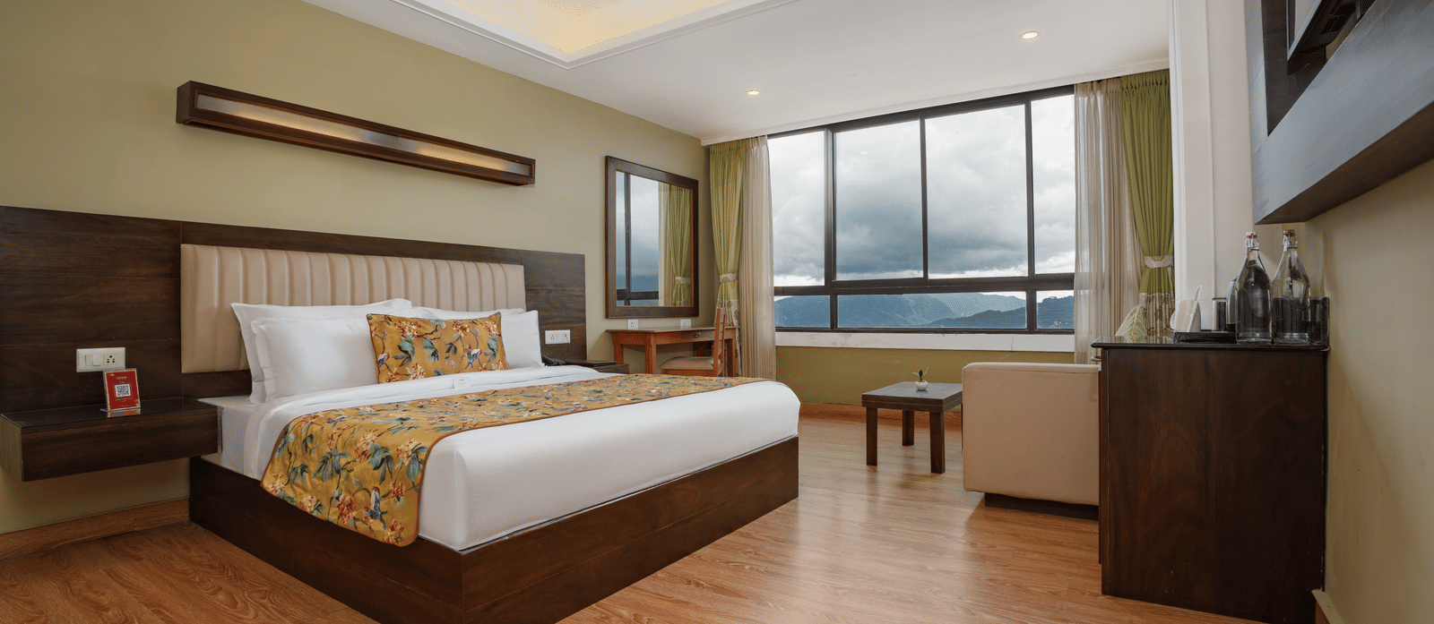 Deluxe Valley View Room at Summit North Star, Gangtok featuring a bed, work desk with chair, sofa chair and table, a cabinet table with tea and coffee maker, and a large window.