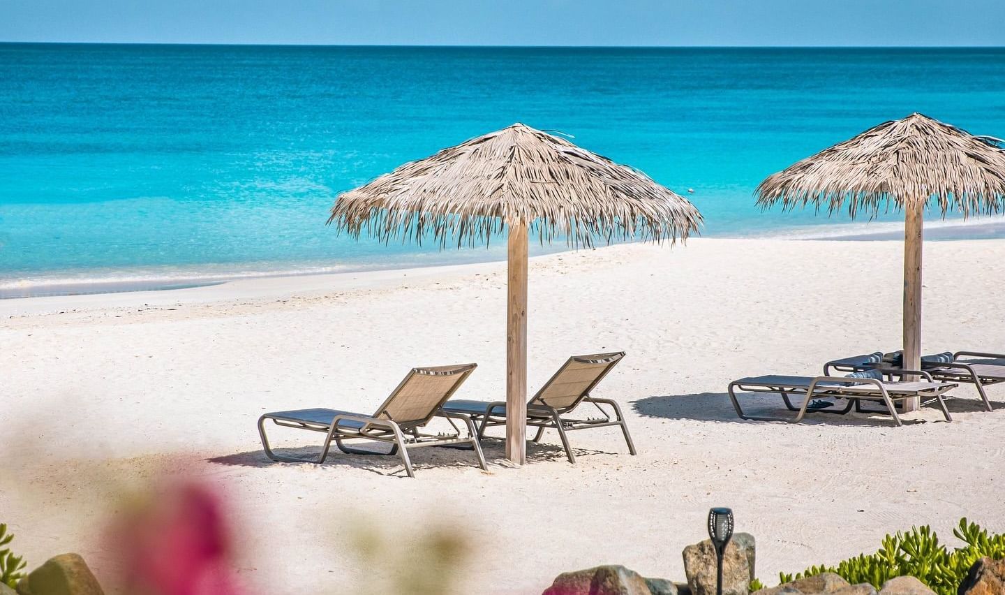 A pristine white sand beach with two thatched umbrellas and lounge chairs, facing a brilliant turquoise ocean.