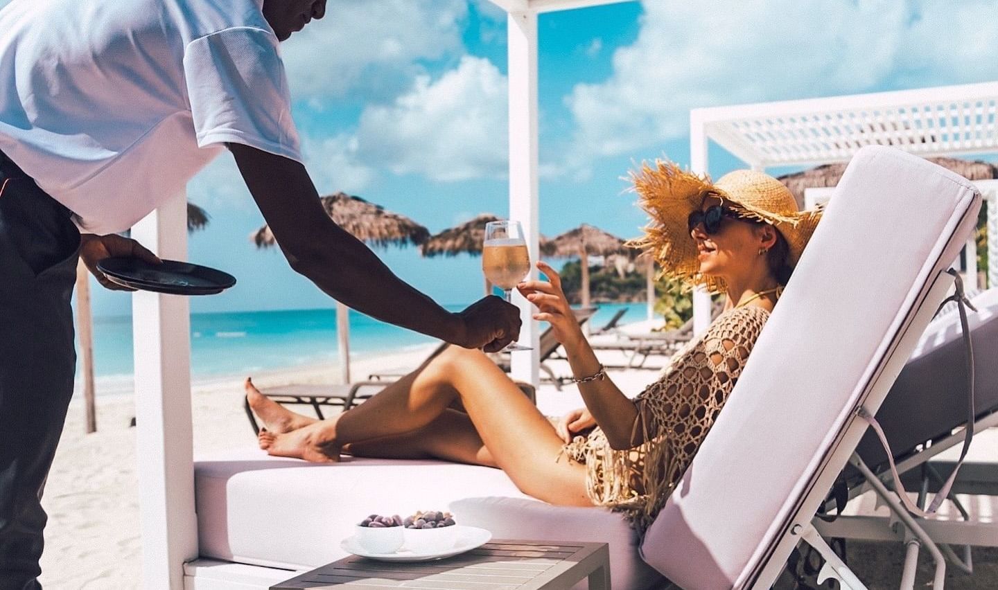 A person serves a drink to a guest relaxing on a lounge chair under a white canopy.
