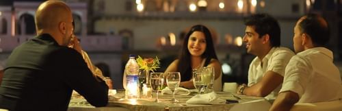 Four people are enjoying an evening meal at a candlelit table outdoors at Tijara Fort-Palace, a Neemrana property in Alwar, with the illuminated fort in the background.