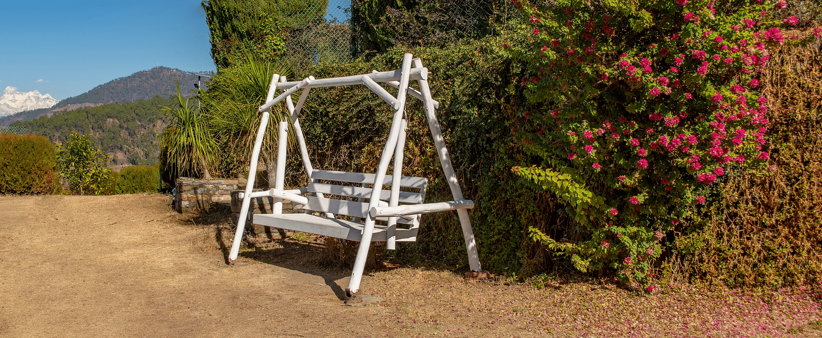 A white wooden swing bench set against vibrant foliage and a rustic garden setting taken from a far angle view. - Blusalzz Homes The Himalayan Bungalow, Almora